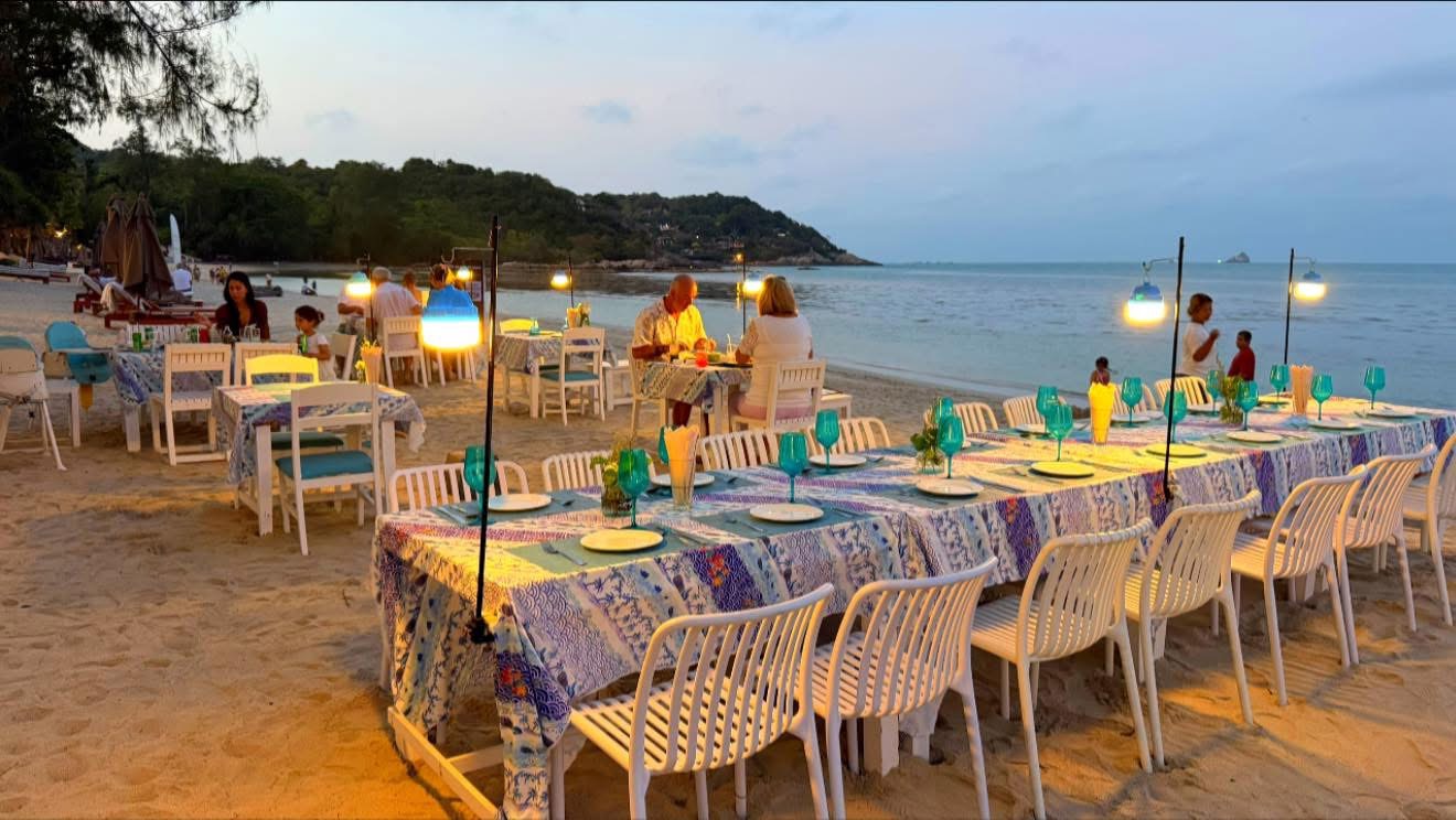 Long tables set on the sand at dusk — blue tablecloths and pendant lights
