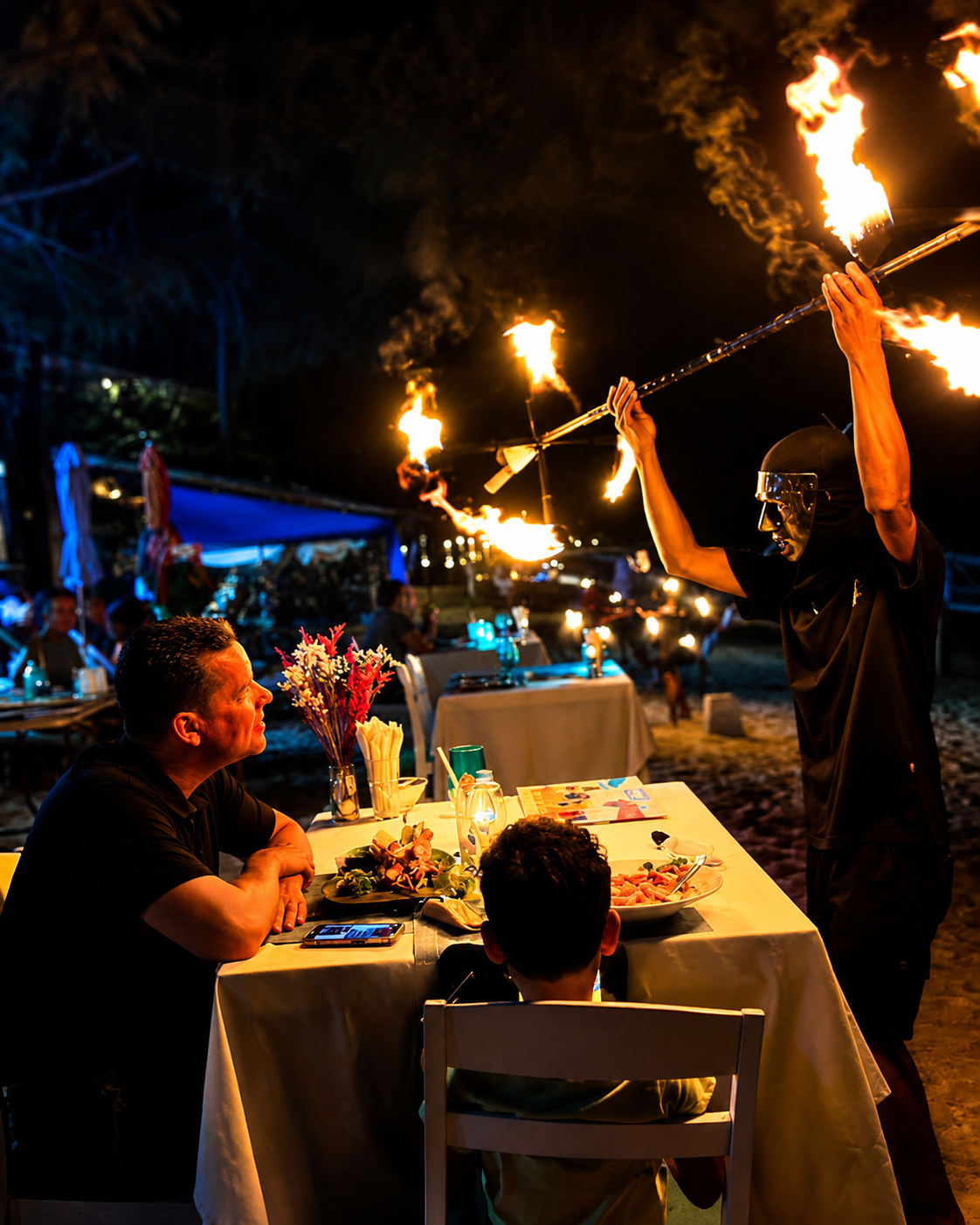 Artista de fuego con bastones en llamas junto a comensales en la playa